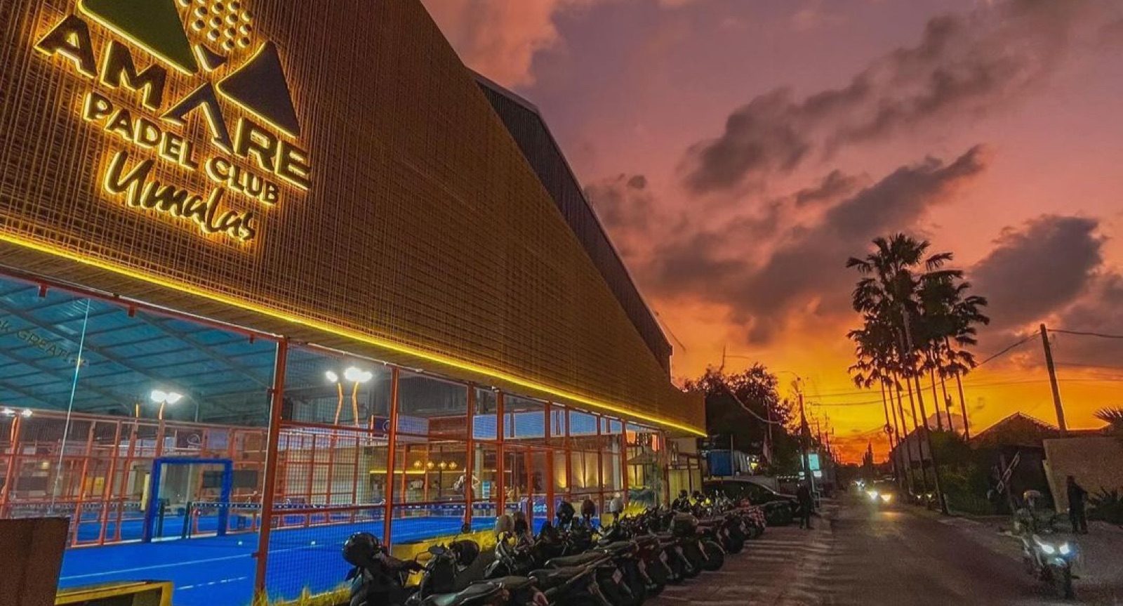 Fasad depan Amare Padel Club Umalas menyala saat golden hour, dengan langit jingga dan siluet pohon kelapa