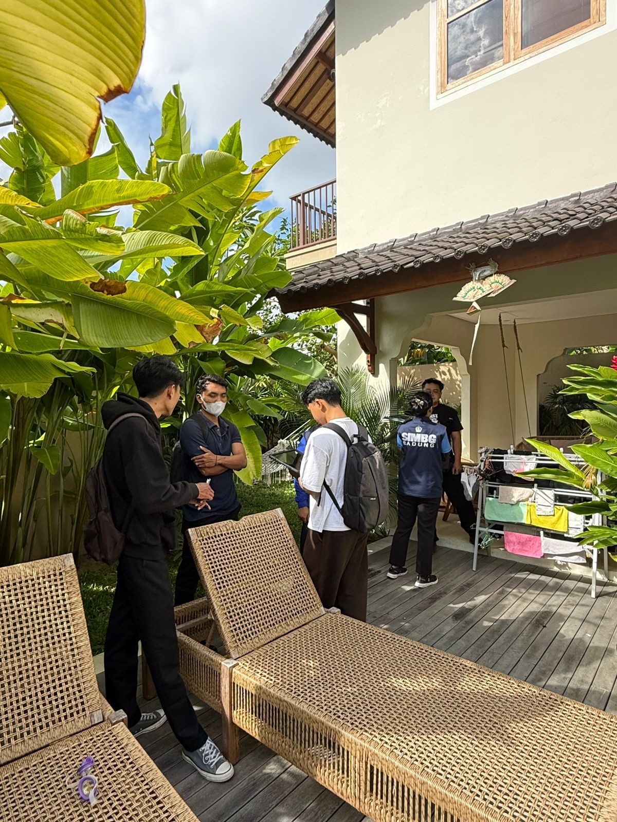 Surveyor berbackpack menyusuri tepi kolam renang menuju bangunan berjendela kaca-kayu besar