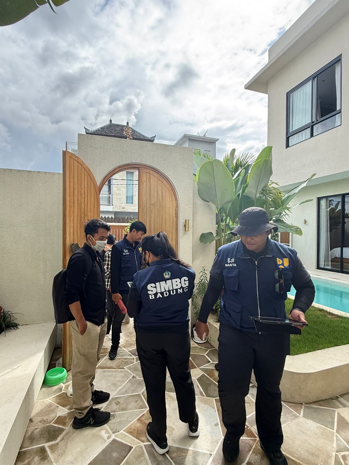 Petugas SIMBG Badung berdiskusi dengan konsultan JKK yang mendampingi di halaman batu alam sebuah villa saat pemeriksaan lapangan, dengan gerbang kayu dan kolam renang di latar belakang