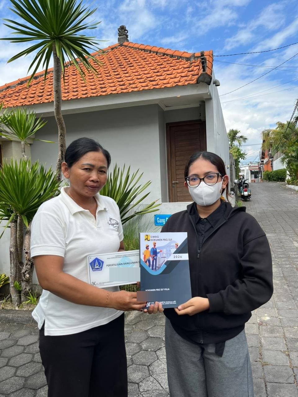 Konsultan JKK menyerahkan plakat Persetujuan Bangunan Gedung kepada klien di depan bangunan di Badung; nomor SK & nama diburamkan